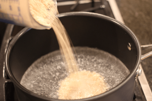 Brown rice being poured into a pot of simmering water on the stove.