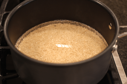 Brown rice added to water in a medium dark pot on the stove before being cooked.