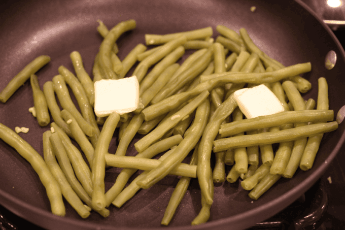 Cooked green beans in a large skillet with a couple pats of butter being melted into them.