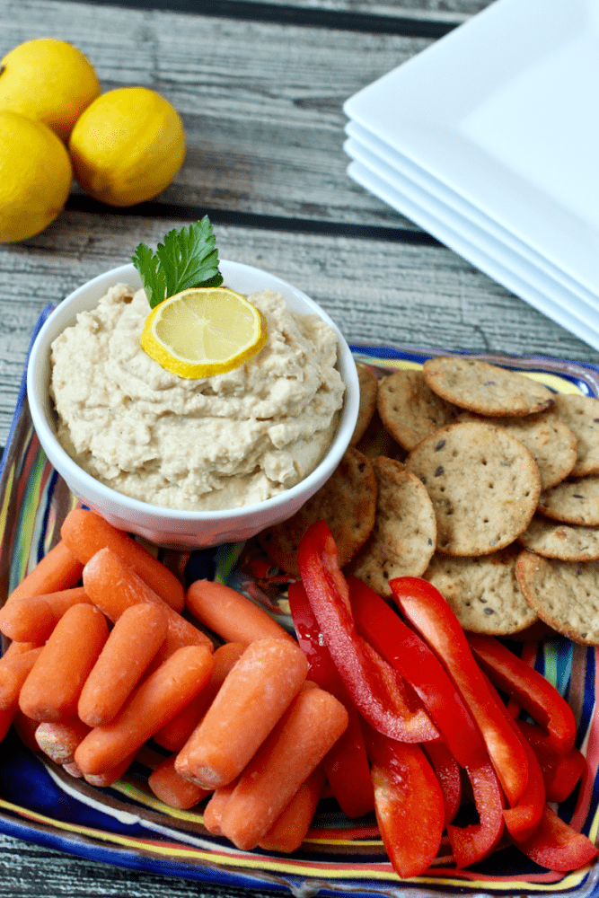 A colorful plate with carrots, red bell pepper strips, crackers and a bowl of homemade hummus with plates in the background for serving