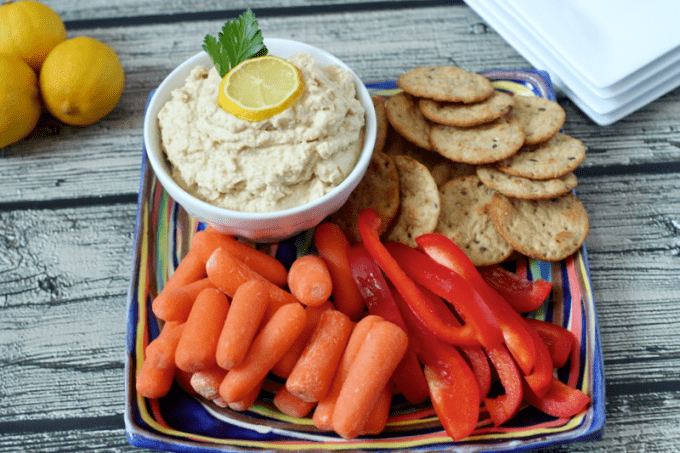 Hummus served with crackers, carrots and bell pepper strips for dipping on a colorful plate.