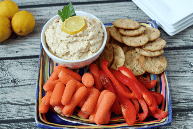 Hummus served with crackers, carrots and bell pepper strips for dipping on a colorful plate.