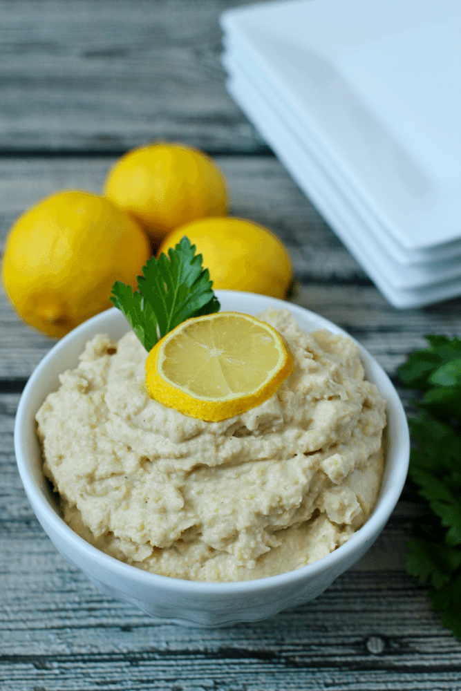 A bowl of healthy homemade hummus in a white bowl with a lemon slice on top and a sprig of parsley, with lemons in the background.