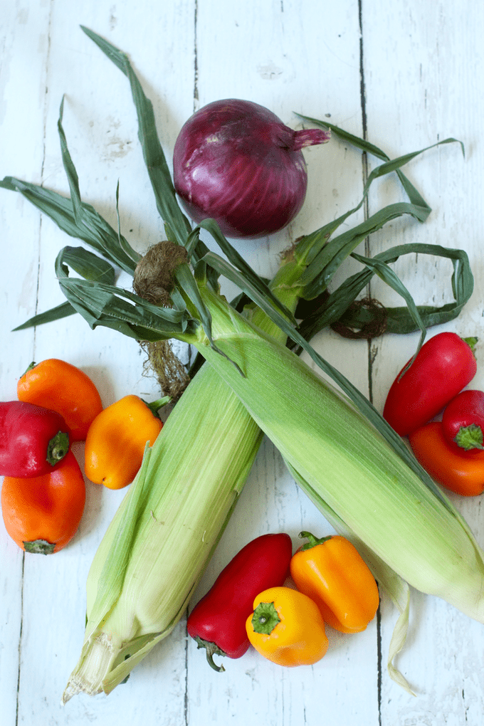 Two ears of corn in their husks along with a mix of mini bell peppers and a red onion