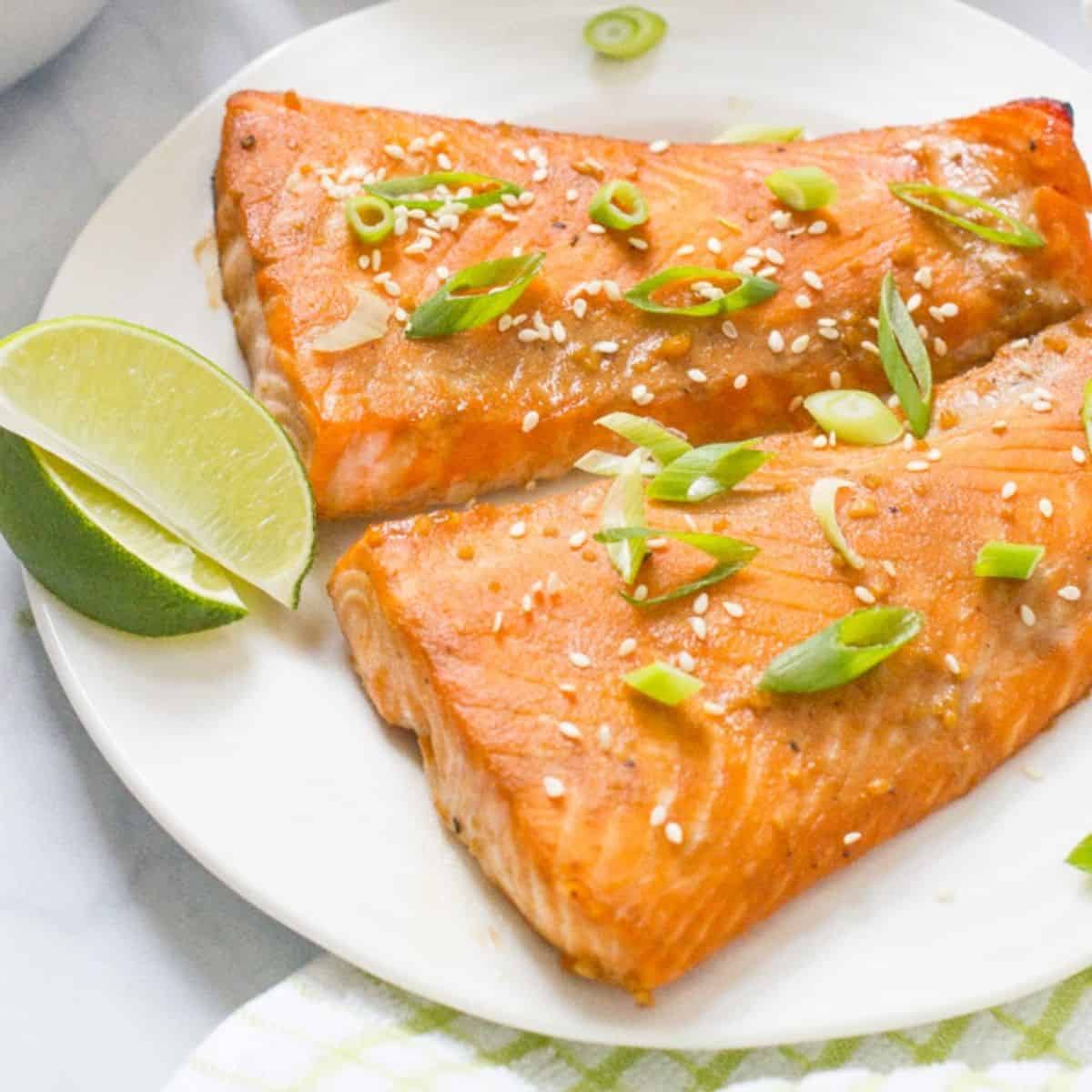 Two cooked salmon filets with a bourbon marinade on a white plate with lme wedges to the side and sesame seeds and green onions sprinkled on top.