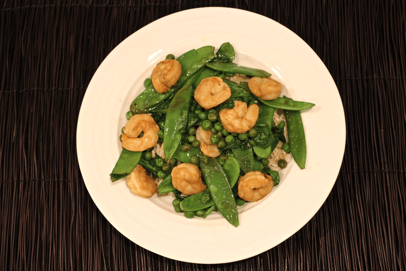 Shrimp stir fry with snow peas, green peas and spinach served in a low white bowl.