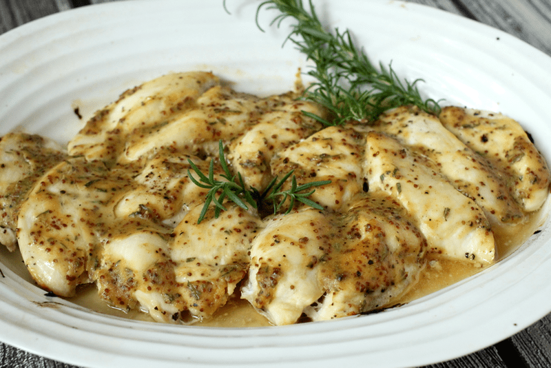 A baked honey Dijon chicken dish with rosemary for garnish in a large white oval baking dish.