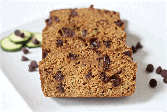 Chocolate chip zucchini bread sliced and served on a white plate with chocolate chips and sliced zucchini coins nearby