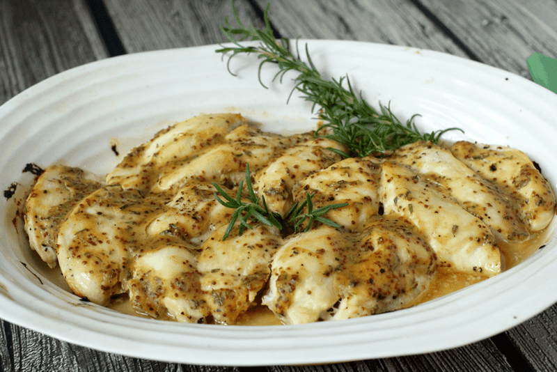 A baked honey Dijon chicken dish with rosemary for garnish in a large white baking dish.