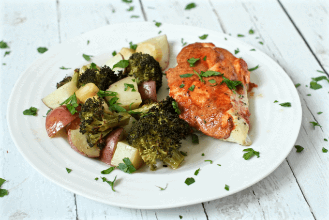 A white dinner plate with roasted potatoes and broccoli alongside a chicken breast with a seasoned mixture baked on top.