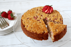 A strawberry cake on a glass cake stand with one piece sliced and being removed.