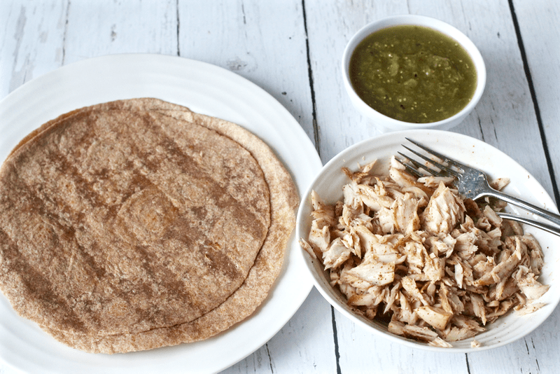 A plate with flour tortillas beside a bowl of seasoned cooked and flaked mahi mahi with a bowl of salsa verde in the background.