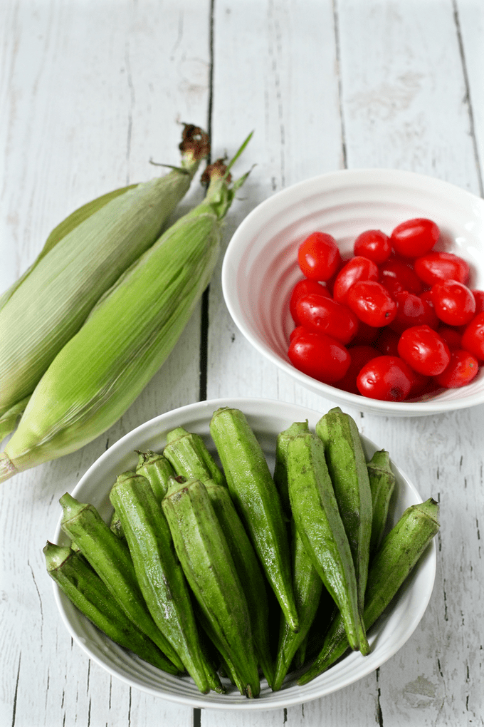 Two ears of corn in the husks, a bowl of cherry tomatoes and a bowl of okra laid out on a white washed counter