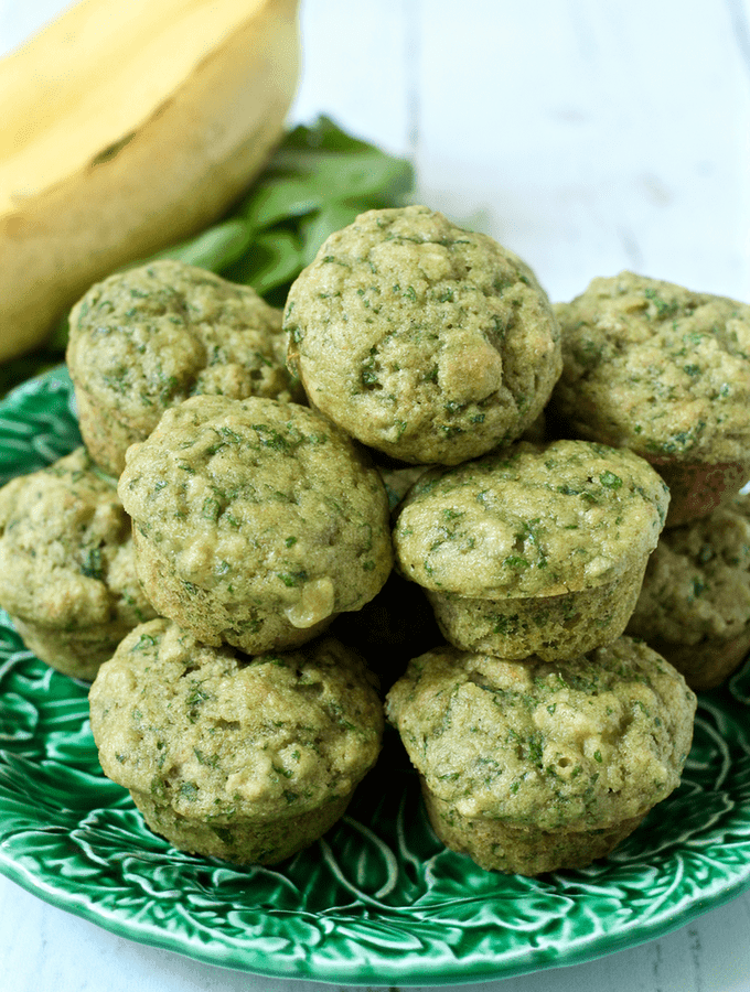A pile of green spinach banana mini muffins on a green leaf plate with fresh spinach and bananas in the background.