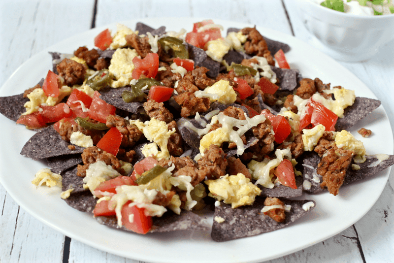 A white plate full of blue tortilla chips topped with scrambled eggs, cooked breakfast sausage, cheese and tomatoes.