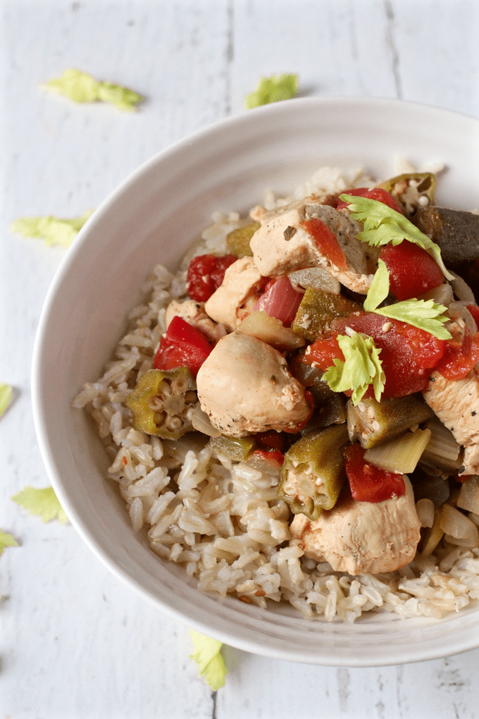 Cajun chicken stew with okra and tomatoes served over rice in a white bowl with celery leaves sprinkled around.
