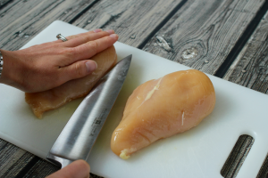 A hand pressing down on the top of a chicken breast while a knife starts to cut it from the side