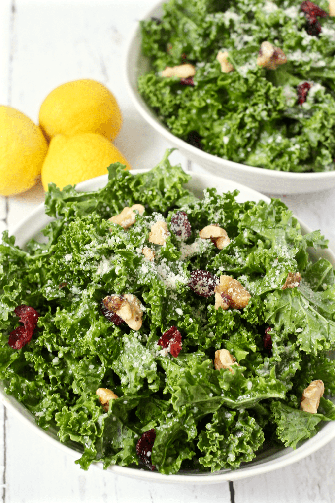 Bowls of a massaged kale salad with walnuts, dried cranberries and Parmesan cheese with lemons to the side.