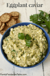 A blue bowl full of a mashed eggplant mixture with fresh parsley on top and crackers to the side.