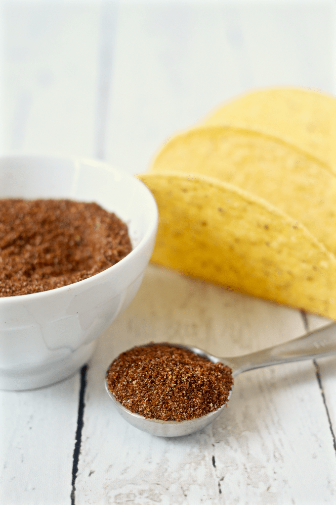 A teaspoon scooped from a bowl of taco seasoning mix with taco shells in the background