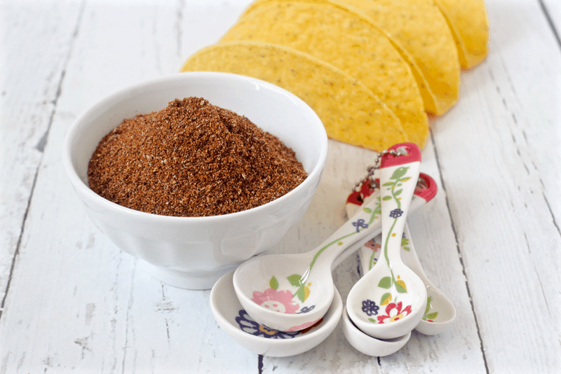 A bowl of homemade taco seasoning alongside measuring spoons with taco shells in the background