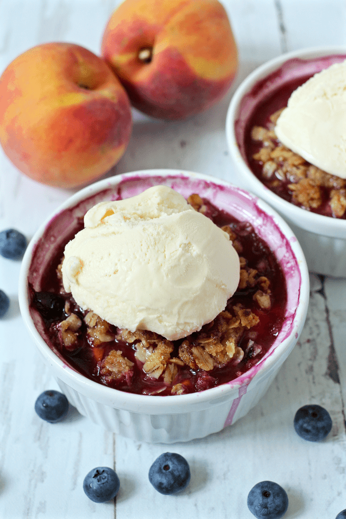 Two baked fruit crumbles in small ramekin dishes with an oat crumble mixture on top and a dollop of vanilla ice cream on each one.