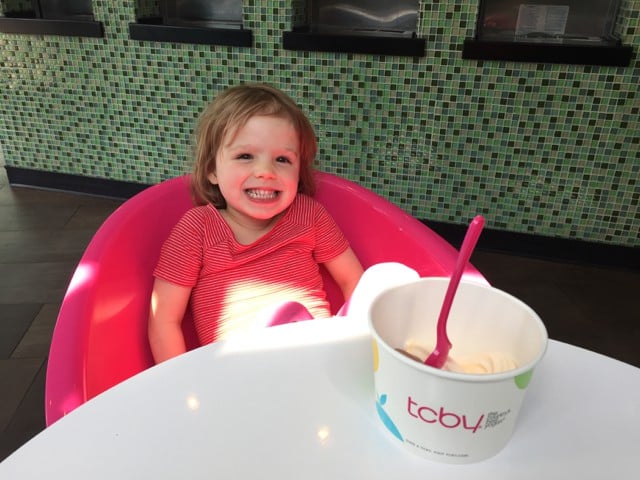 A young girl in a big pink plastic chair with a cup of TCBY frozen yogurt on the table in front of her.