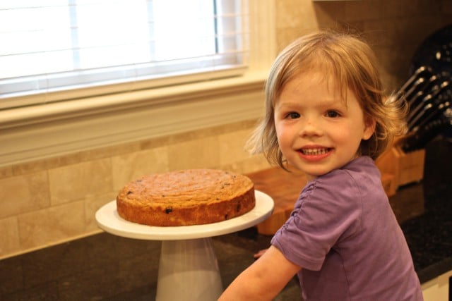 A young girl in a purple shirt next to a white cake stand with a small baked cake on it.