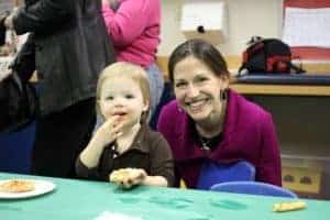 A woman and her small child who is seated at a little table and eating an English muffin pizza.