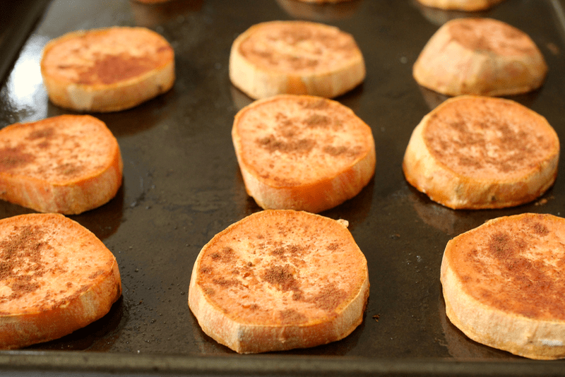 Sliced sweet potato rounds sprinkled with cinnamon lined up on a baking sheet  before being cooked.