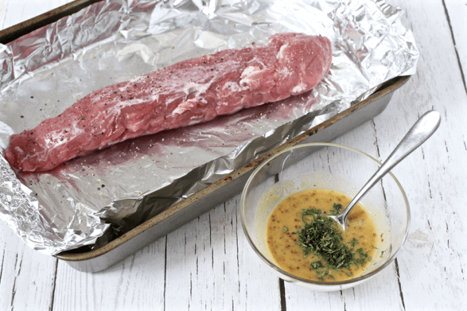 A trimmed and seasoned raw pork tenderloin on a foil lined baking sheet with a bowl of a mustard rosemary sauce to the side.