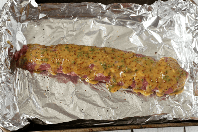 A foil lined baking sheet with a pork tenderloin topped with a maple mustard sauce with rosemary.