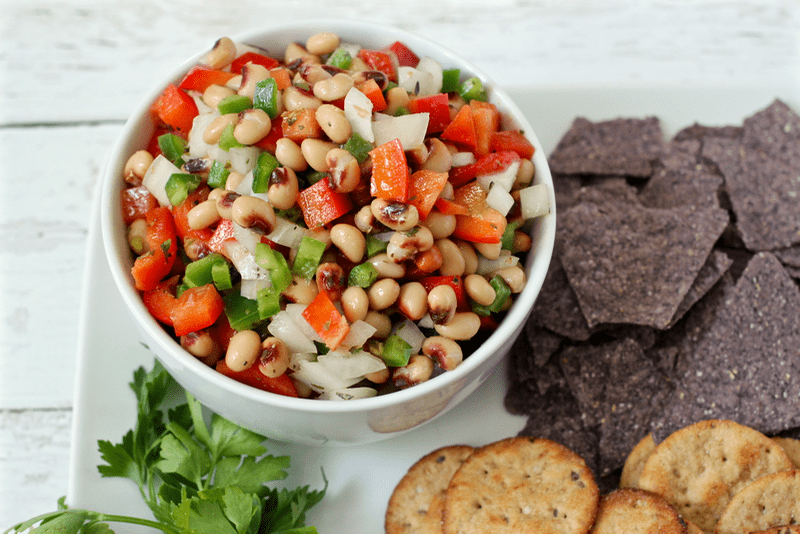 A black eyed pea dip in a white bowl on a platter with tortilla chips and crackers for dipping.