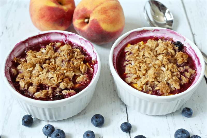 Two baked peach and blueberry crumbles in small ramekin dishes with an oat crumble mixture on top and additional fresh fruit to the side.