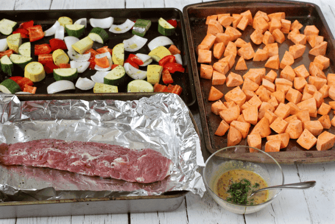 Separate baking pans laid out, one with cubed sweet potatoes, one with a mix of chopped onions, zucchini, squash and red bell pepper and one with a pork tenderloin.