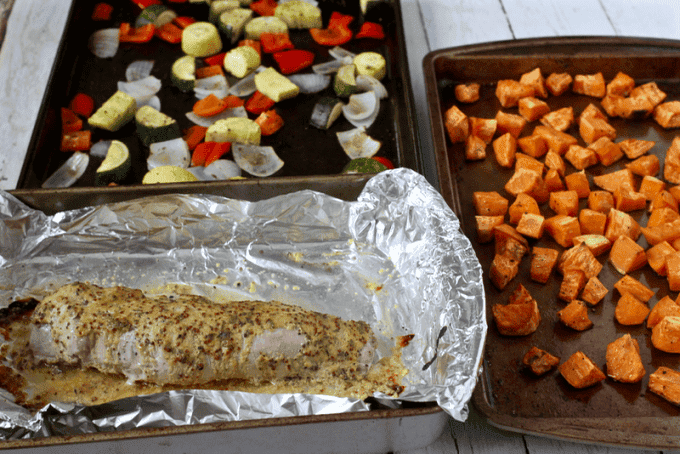Separate baking pans laid out, one with roasted sweet potatoes, one with a mix of roasted onions, zucchini, squash and red bell pepper and one with a pork tenderloin in a mustard sauce.