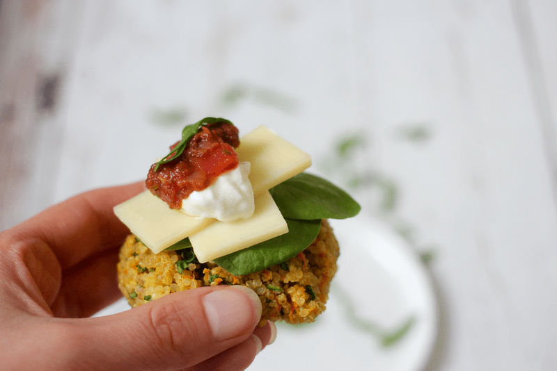 A hand holding up a mini quinoa veggie patty topped with spinach, sliced cheese, sour cream and salsa.