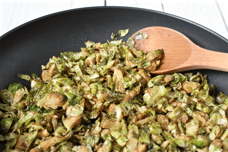 A wooden spoon resting in a skillet of sautéed shredded Brussels sprouts with balsamic vinegar.