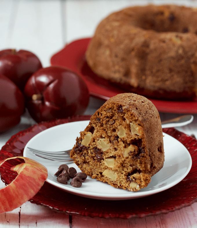 A tall slice of an apple cake with chocolate chips on a white plate set on a red plate with the remaining cake in the background.