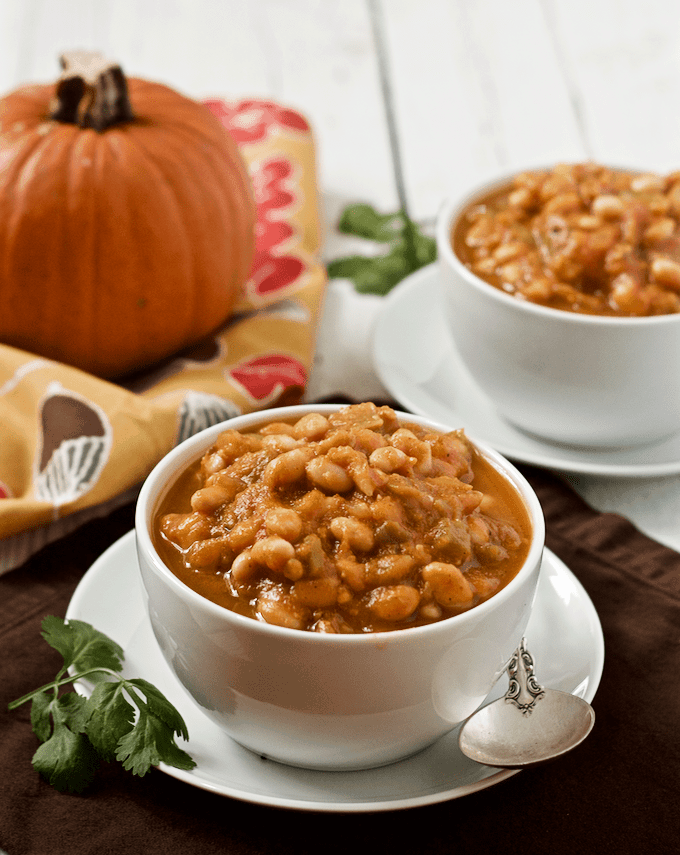A pumpkin white bean chili served in two white bowls set on white plates and a small pumpkin in the background.