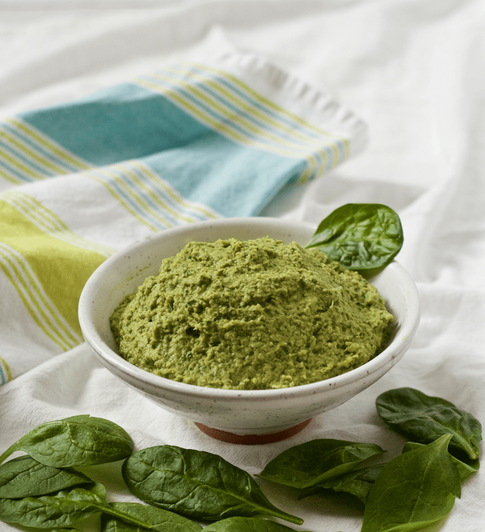 Spinach artichoke hummus served in a white bowl with leaves of fresh spinach scattered nearby