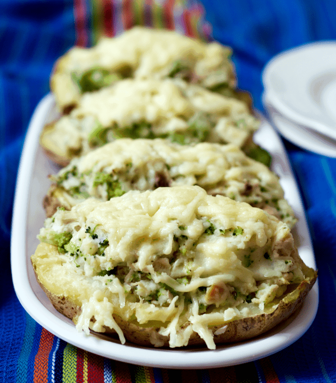 Four baked potato halves stuffed with a cheesy chicken and broccoli mixture and topped with melted white cheddar cheese and served on a long oval platter.