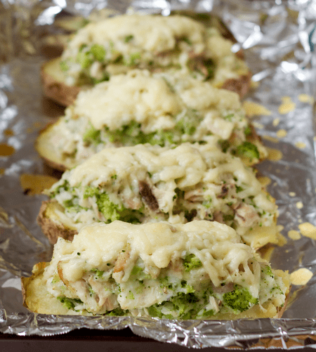 Four baked potato halves stuffed with a cheesy chicken and broccoli mixture and topped with melted white cheddar cheese on a foil lined baking sheet.