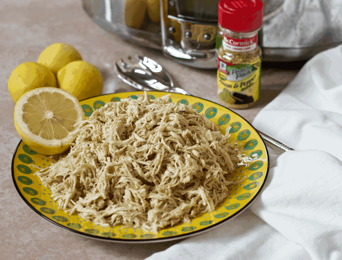 Shredded chicken piled on a yellow plate next to a slow cooker and a spice container of lemon pepper seasoning.