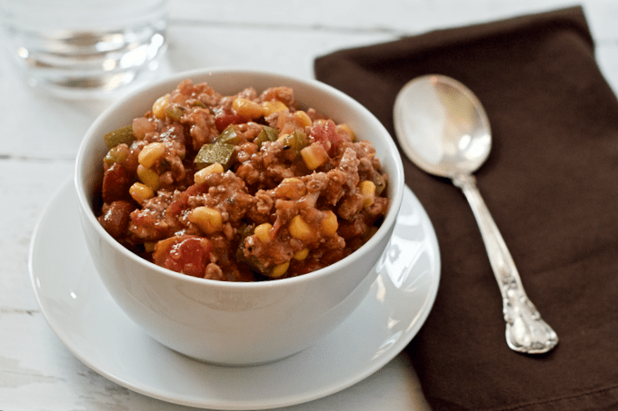 A bowl of a ground turkey, bean, corn and veggie stew in a white bowl alongside a brown napkin with a spoon on top.