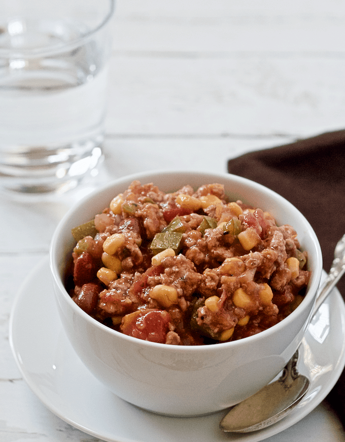 A white bowl set on a plate and filled with a ground turkey stew with beans, corn and veggies.