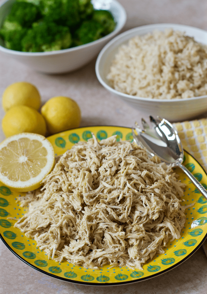 Shredded lemon pepper flavored chicken piled on a yellow plate with bowls of rice and broccoli in the background.