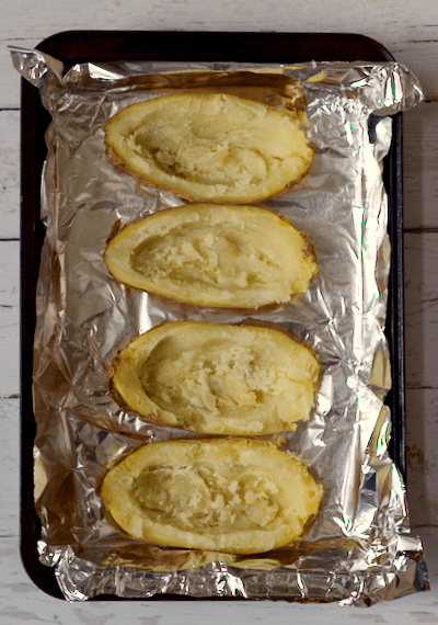Four baked potato halves scooped out and on a foil lined baking sheet before being stuffed.