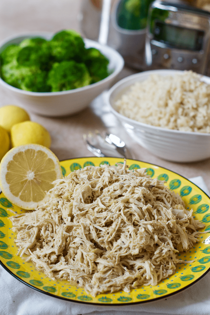 Lemon pepper shredded chicken on a yellow plate with bowls of broccoli and rice in the background next to a slow cooker.