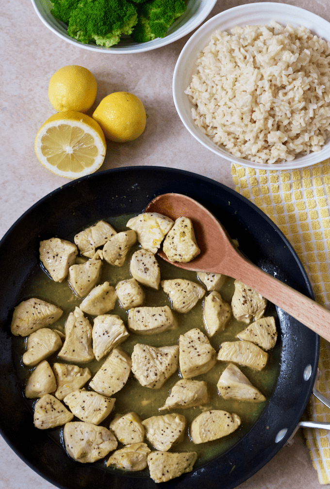 A dark skillet with cubed cooked chicken seasoned with a lemon pepper seasoning with a bowl of rice and lemons to the side.